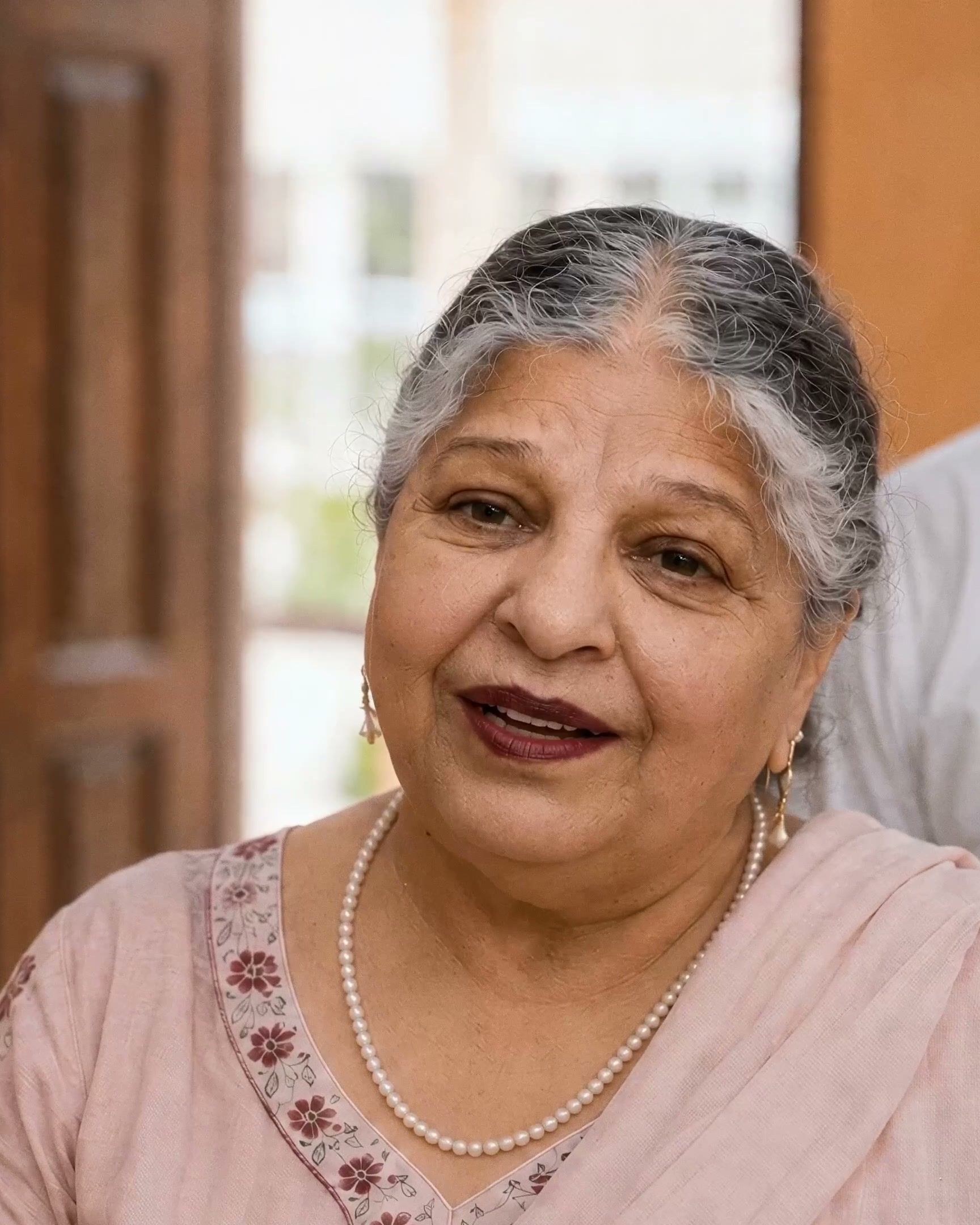 An elderly woman in an embroidered kurta and pearls smiles to camera in soft daylight.